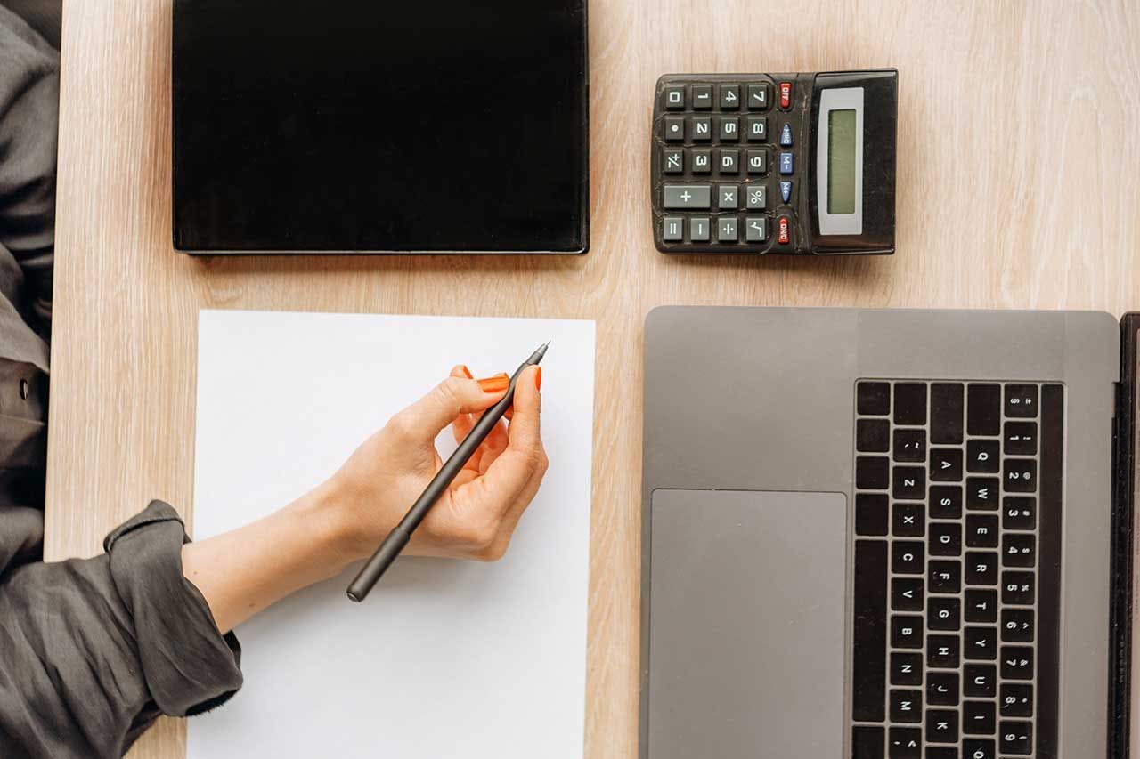 Eine Person berechnet die Rendite einer Immobilie aus, in dem sie Blattpapier, Stift, Taschenrechner, Notizbuch und einen Laptop verwendet.